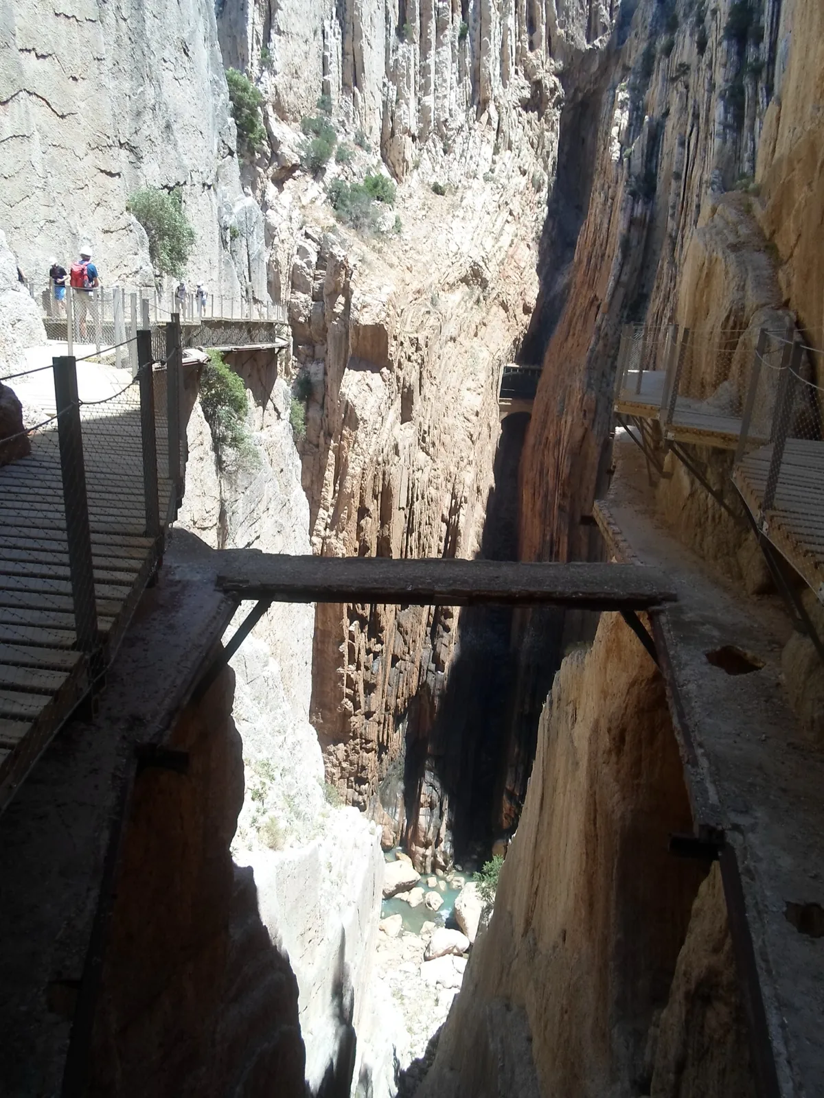 A deep, narrow section of the Gaitanes Gorge where new boardwalks thread along both cliff walls and a crumbling original concrete ledge spans the gap, the river far below