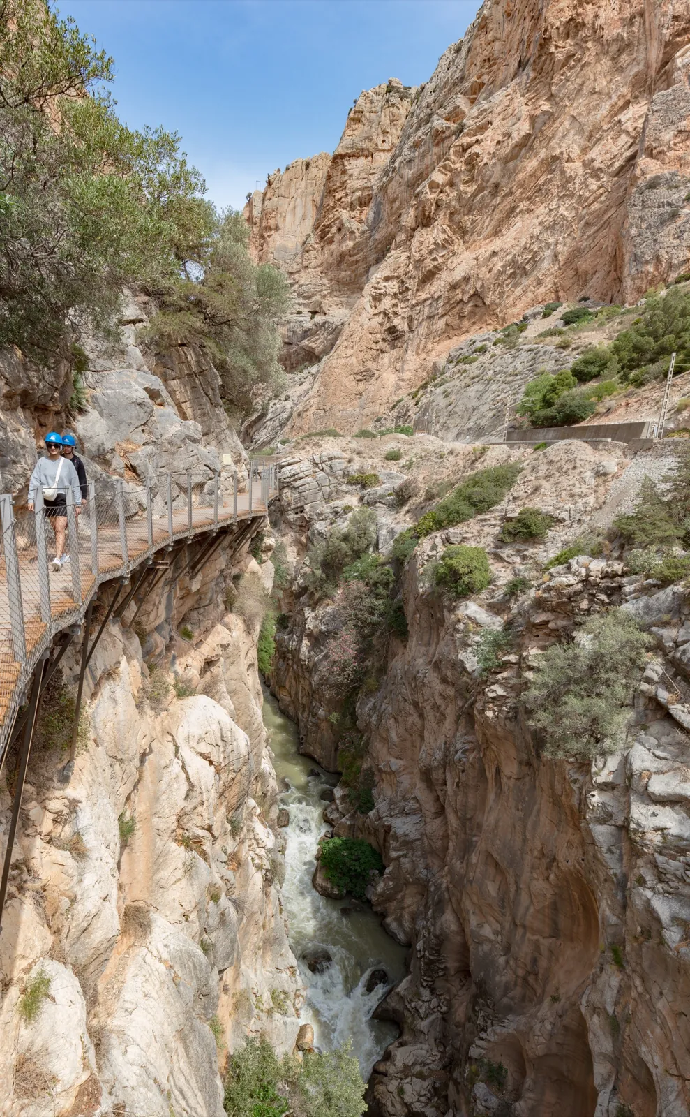 A walker in a blue safety helmet on the narrow railed boardwalk of the Caminito del Rey, clinging high to a limestone cliff with the green Guadalhorce river rushing far below